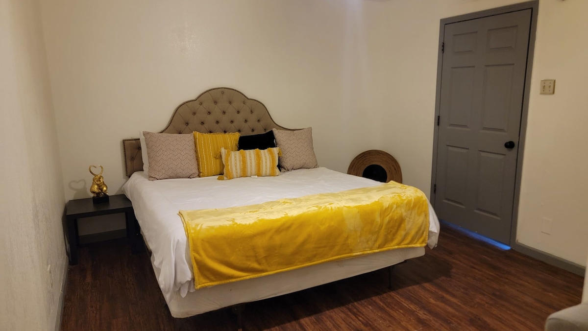 A well-appointed guest suite features a spacious bed adorned with yellow accents and neutral pillows. A bedside table is placed beside the bed, complementing the minimalist decor. Natural light enters from a nearby door, enhancing the inviting ambiance of the room.