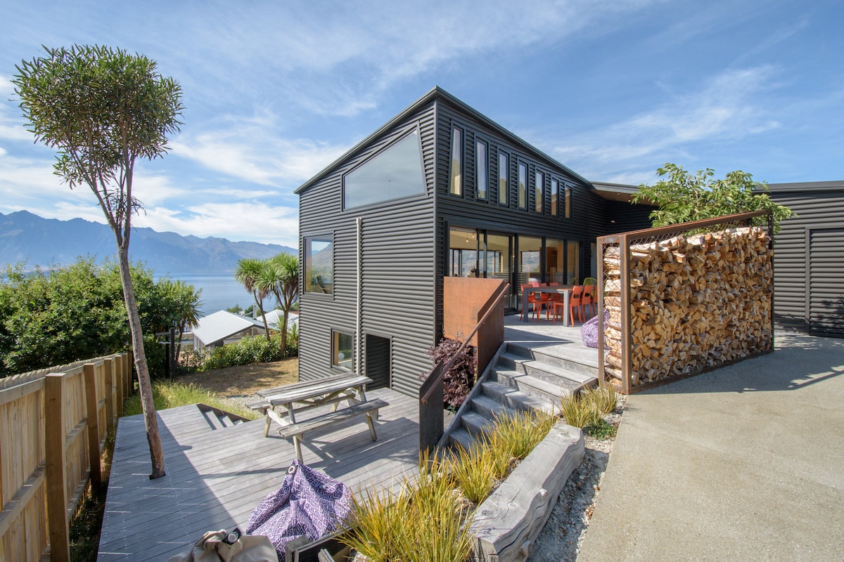 The modern architectural home is presented with a sleek exterior and generous windows. A wooden picnic table is situated on a deck with steps leading to a terraced area. Surrounding greenery and lake views complement the scene, all set against a backdrop of the Remarkables mountains.
