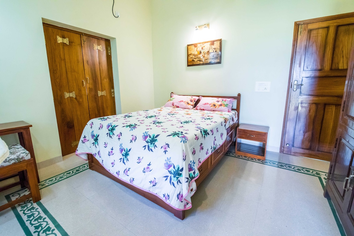 A bright bedroom features a spacious bed adorned with a floral-patterned coverlet. Wooden furniture, including a bedside table, complements the room's light green walls and natural wooden doors. A piece of wall art adds a serene touch, while light filters in, enhancing the inviting atmosphere.