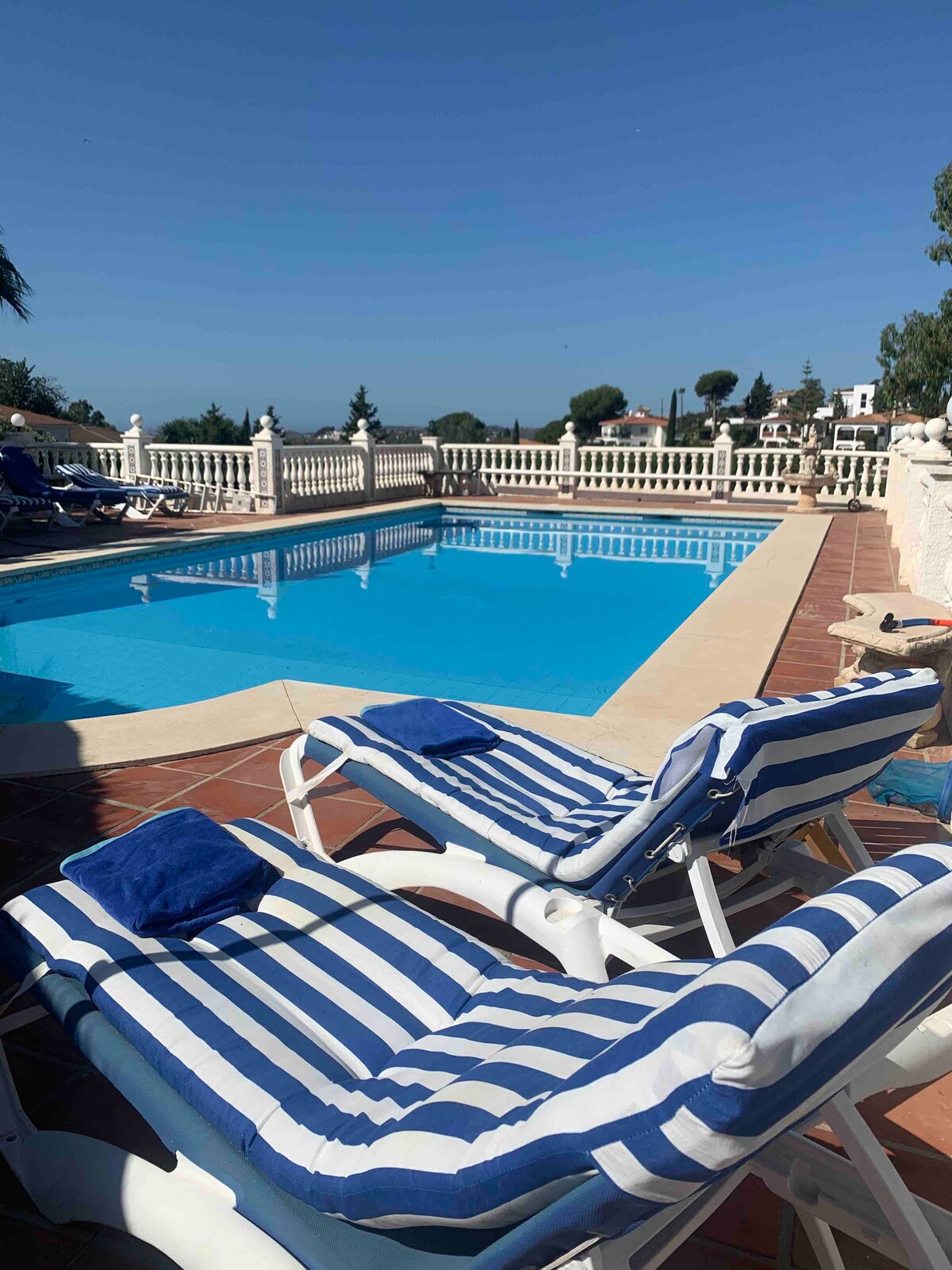 Two striped lounge chairs are positioned beside a crystal-clear swimming pool. The pool area is surrounded by a white railing and offers a panoramic view of the landscape. Bright blue skies are visible overhead, enhancing the outdoor ambiance.