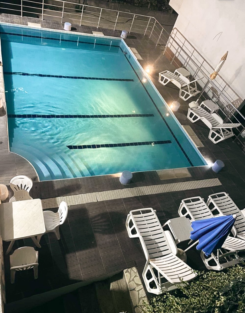 An outdoor swimming pool is displayed, featuring a clear blue water surface illuminated by surrounding lights. Several lounge chairs are arranged poolside, accompanied by a table and an umbrella. The area is neatly paved, offering a comfortable space for relaxation.