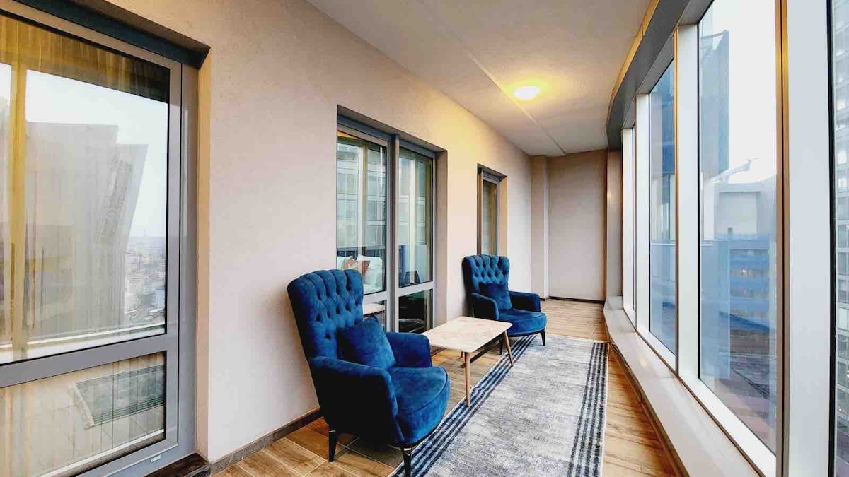 A sunlit terrace features two plush blue chairs set around a small coffee table, inviting relaxation. Large windows provide expansive views of the surroundings, and a neutral rug adds a touch of warmth to the space, which is enhances by soft natural light.