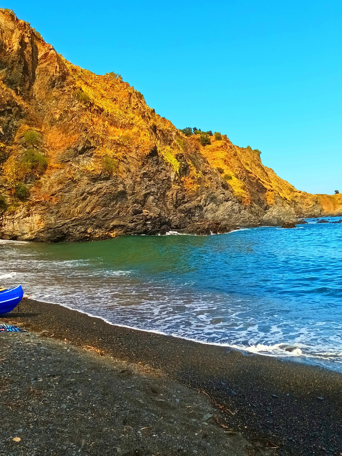 A rocky coastline is depicted, with golden cliffs rising above a tranquil blue sea. The water gently laps at a pebbled shore, reflecting the clear blue sky. The natural landscape conveys a serene environment inviting exploration.