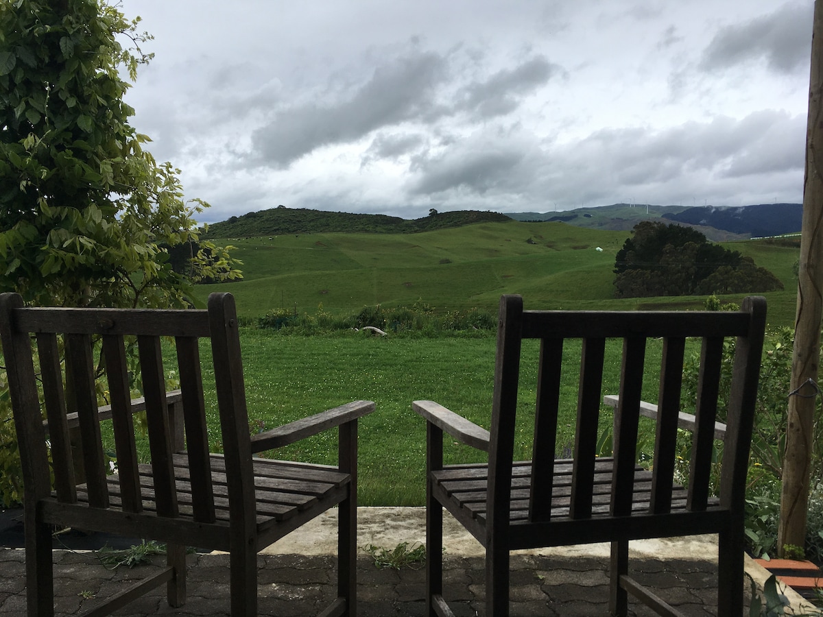 Two wooden chairs are positioned on a patio, providing a clear view of the expansive green meadows and rolling hills in the background. Cloudy skies create a tranquil atmosphere, highlighting the natural surroundings.