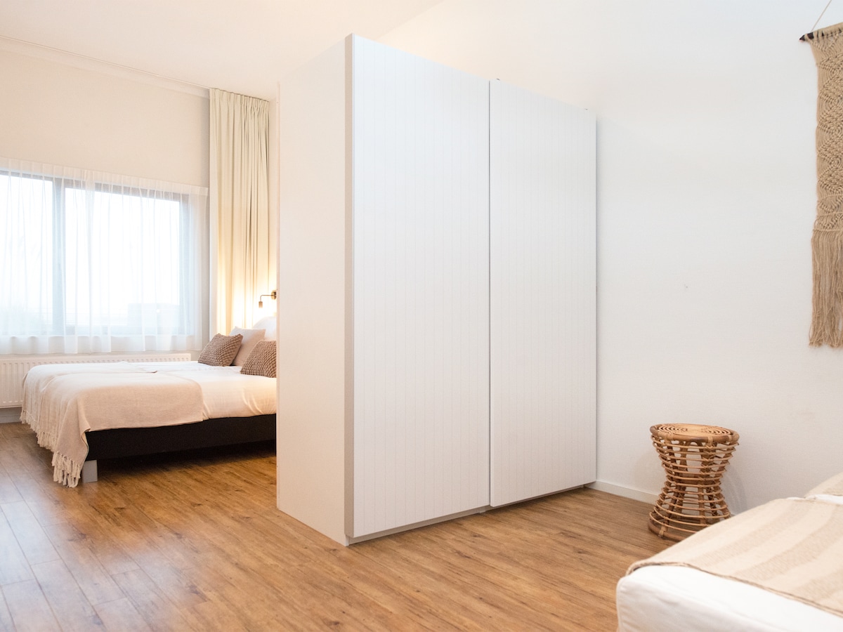 The image shows a well-lit bedroom featuring two spacious beds, arranged alongside a wardrobe with sliding doors. Natural light enters through large, sheer curtains, enhancing the warm wood tones of the floor. A small side table constructed of woven material is positioned nearby.