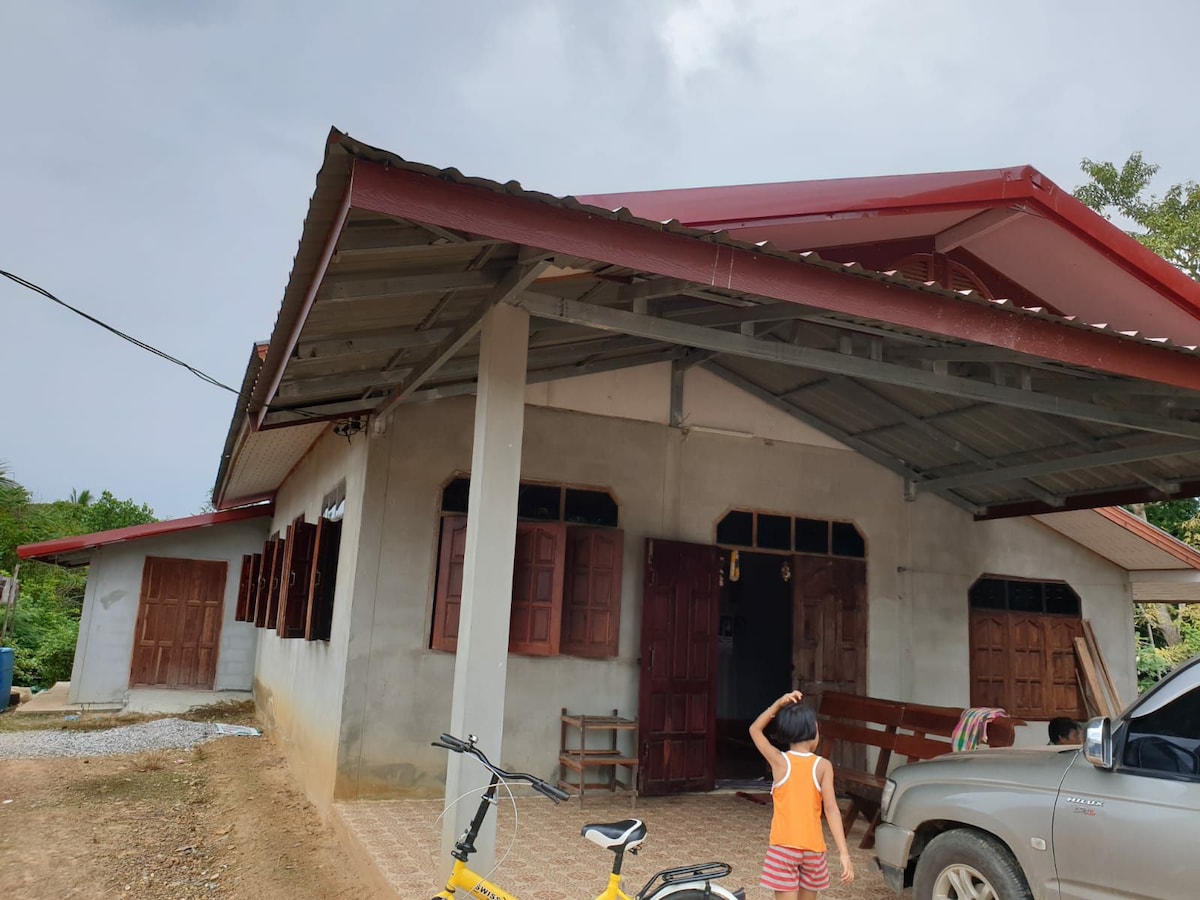 A single-story house features a red metal roof and a concrete exterior, with wooden windows creating a rustic look. A porch extends across the front, and a vehicle is parked nearby, while a bicycle is positioned to one side. Greenery is visible in the background.