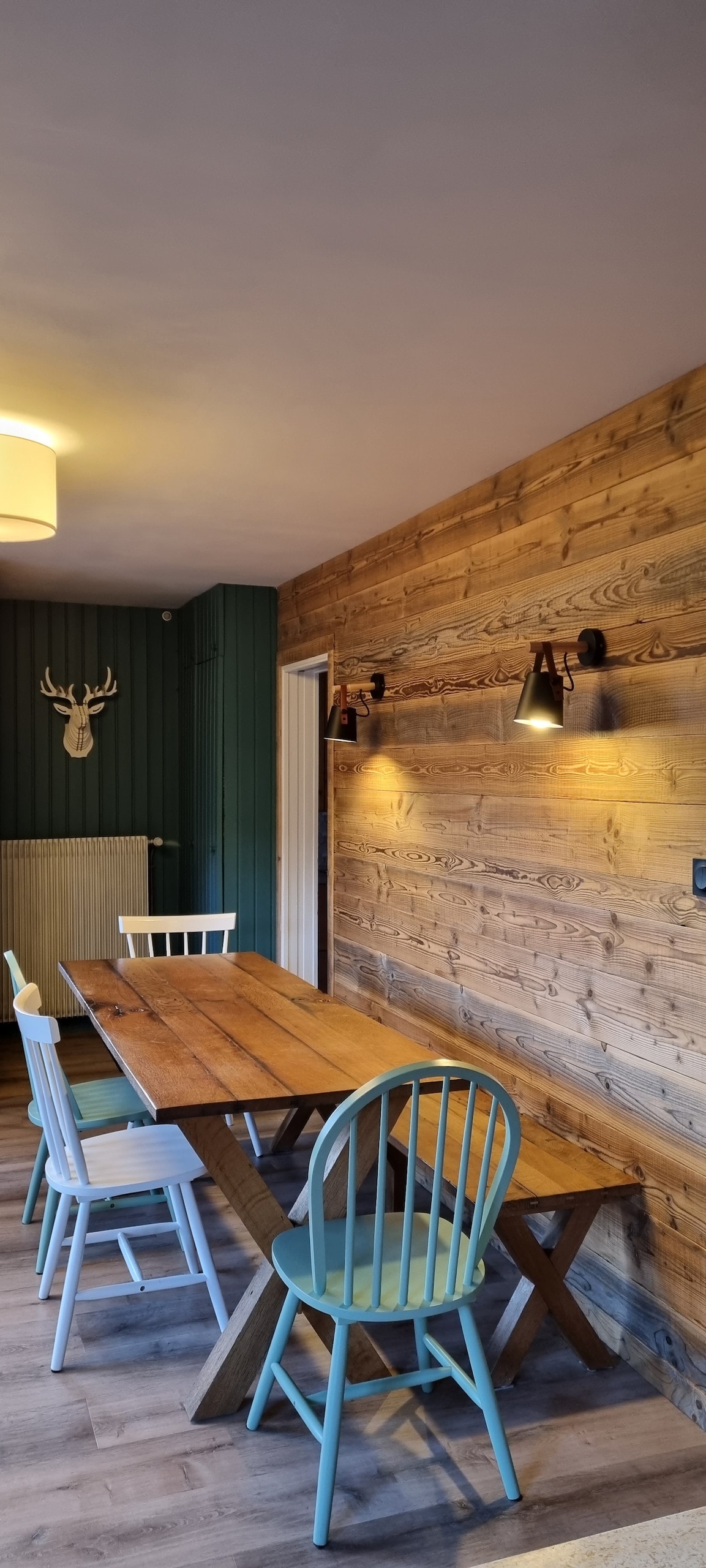 A wooden dining table is set in a cozy dining area, surrounded by chairs in varied styles. The wall features natural wood paneling, with modern light fixtures enhancing the space. A subtle decorative element is present on the wall, contributing to the chalet's inviting ambiance.