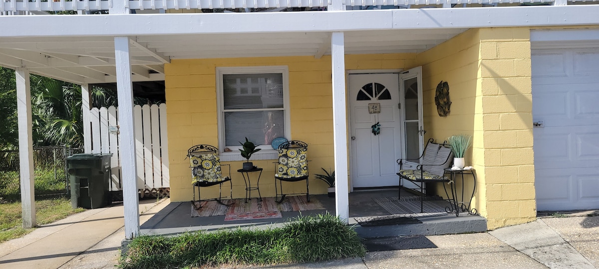 A welcoming front porch features two patterned chairs positioned beside a small table. A decorative door with a circular window stands as the main entry. Light yellow exterior walls provide a bright backdrop, while a small rug adds comfort to the outdoor space.