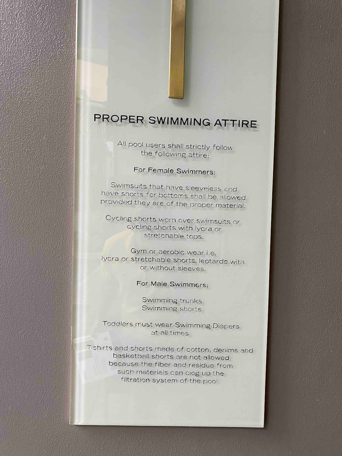 A signage displayed on a wall outlines proper swimming attire for pool users. The text details acceptable clothing for female and male swimmers, including guidelines on swimming trunks and attire suitable for toddlers.