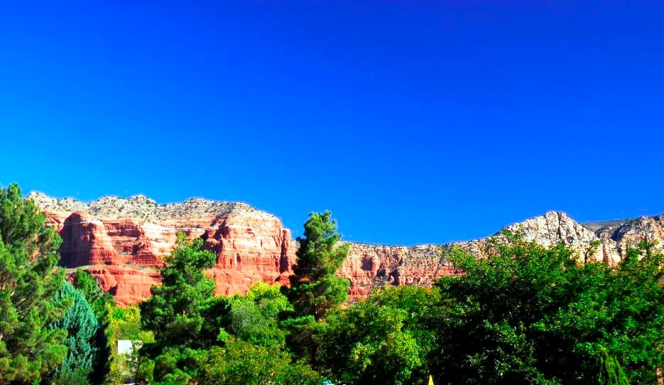 A vibrant landscape is displayed, featuring stunning red rock formations under a clear blue sky. Lush greenery is seen in the foreground, providing a contrast to the rocky terrain of the mountains in the background.