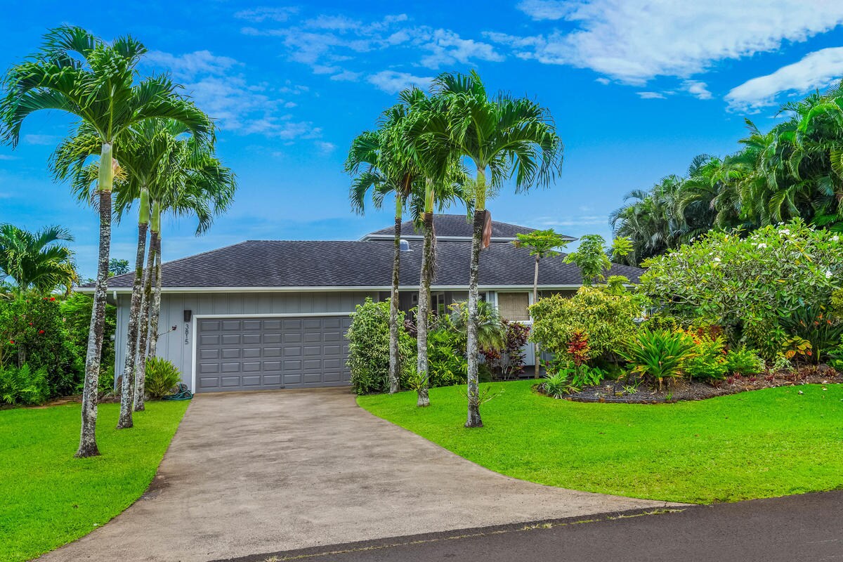 A single-story home is surrounded by lush tropical landscaping, featuring palm trees and colorful plants. The driveway leads to a two-car garage, with a well-maintained lawn providing ample green space. A clear blue sky enhances the serene ambiance of the property.