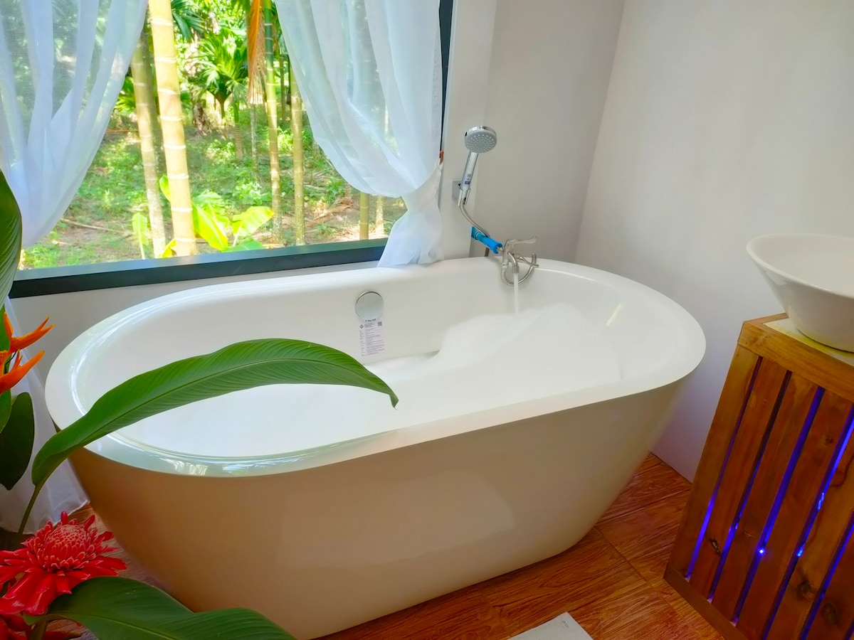 A freestanding bathtub is positioned beside a large window, offering views of greenery outside. Sheer white curtains frame the window, allowing natural light to filter in. A wooden stand complements the serene setting, enhancing the tranquil atmosphere of the bathroom.