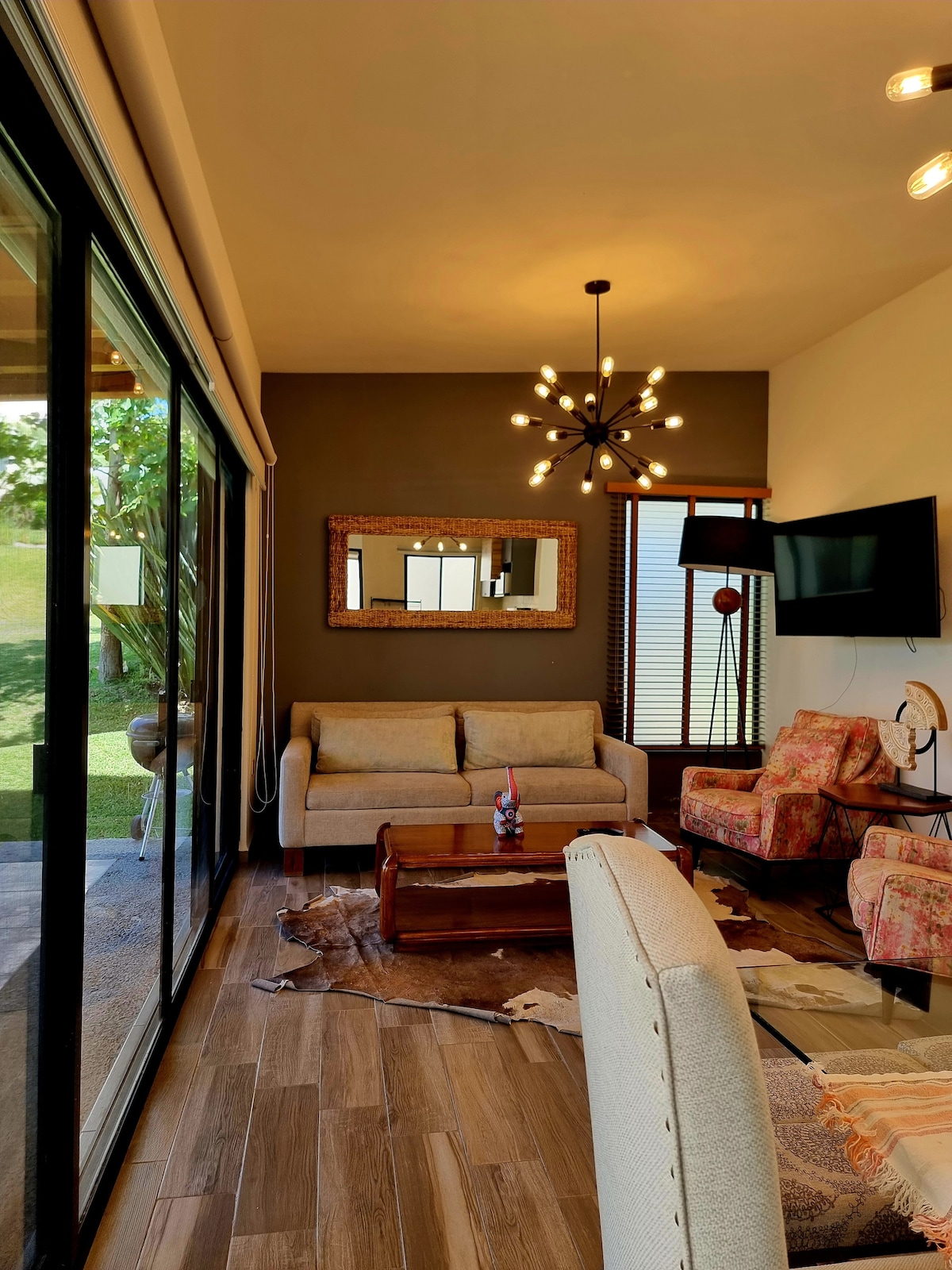 A cozy living area features large glass doors allowing natural light to fill the space. A comfortable gray sofa is complemented by two patterned armchairs. A wooden coffee table centers the room, while a decorative mirror and a modern light fixture add to the ambiance.