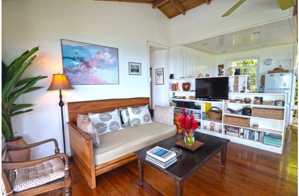 A cozy living area is displayed, featuring a wooden sofa with patterned cushions and a wooden coffee table. A small lamp illuminates the space, while a vibrant floral arrangement adds a pop of color. A wall-mounted TV and well-organized shelves are visible in the background.