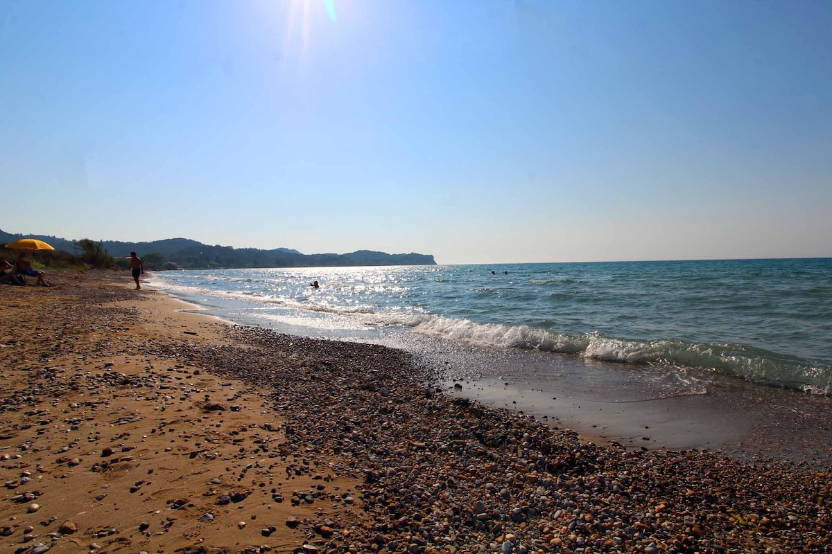 The image showcases a serene beach scene with gentle waves lapping at the pebbled shore. Sunlight reflects off the water, creating a shimmering effect. The coastline features soft hills in the background, and a few sun loungers are visible along the beach.