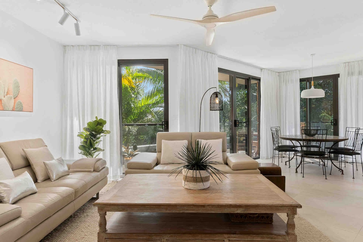 A modern living area features a light-colored leather sofa and a wooden coffee table with a decorative plant. Large windows with sheer curtains allow natural light to illuminate the space, revealing a view of palm trees. A dining area with a round table and black chairs is visible in the background.