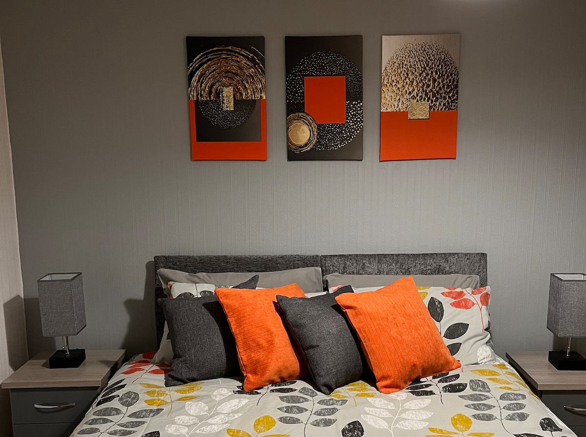 A comfortable sleeping area features a sleek bed adorned with decorative cushions in vibrant orange and gray. Stylish bedside lamps flank the bed, while abstract wall art in coordinating colors adds visual interest above the headboard.