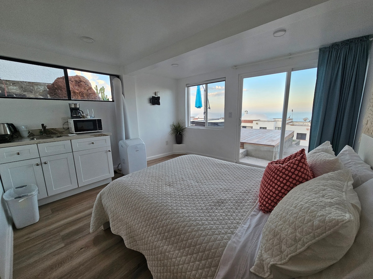 A bright sleeping area is presented with a spacious bed adorned with textured bedding and decorative pillows. Large windows provide a view of the outdoors, while a kitchenette with essential appliances is situated nearby, contributing to the functional design.