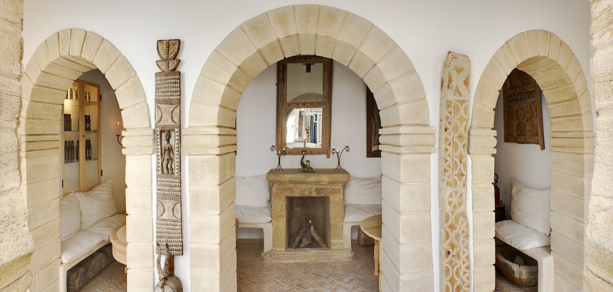 A welcoming seating area is framed by three elegant stone arches. The space features a simple stone fireplace, surrounded by comfortable, plush seating. Natural light fills the room, highlighting the warm tones of the decor and the textured walls.