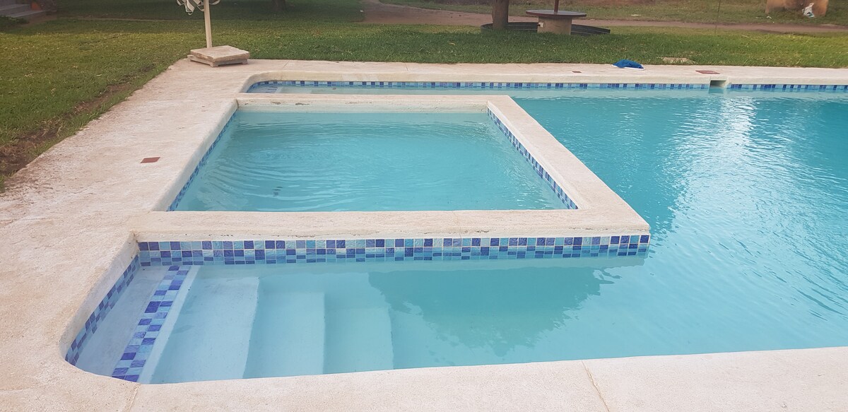 A swimming pool area is shown with a spacious rectangular pool featuring clear blue water. A smaller attached hot tub is visible at one end, with steps leading into the pool. Surrounding grass provides a natural setting.