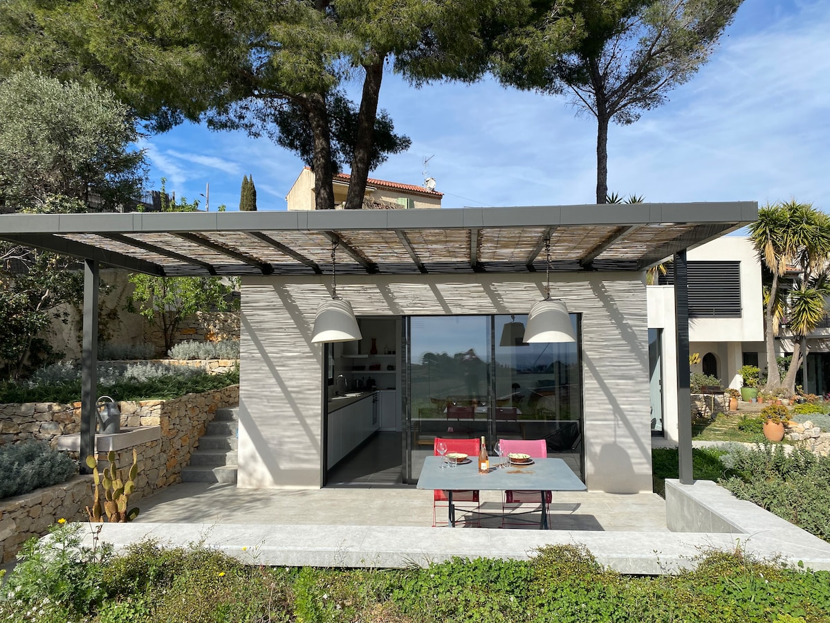 An architect-designed extension showcases a modern exterior with large windows. A shaded patio features a dining table, accompanied by two hanging pendant lights. Surrounding greenery adds to the serene atmosphere, while the distant view highlights the natural beauty of the landscape.