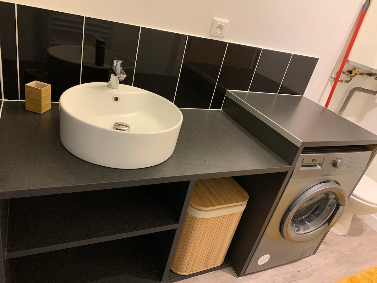 A modern bathroom space features a circular sink atop a sleek black countertop, complemented by wall tiles in a contrasting pattern. A washing machine is positioned beside a wooden waste bin, adding functional elements to the overall design.