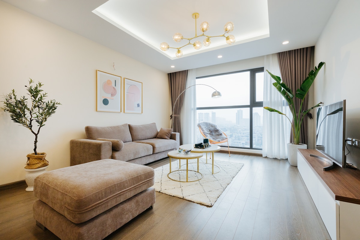A spacious living area features a large, plush sofa alongside a matching ottoman. A decorative plant adds a natural touch, while modern artwork adorns the walls. Large windows provide ample light and stunning city views, complemented by cozy textiles on a soft rug.