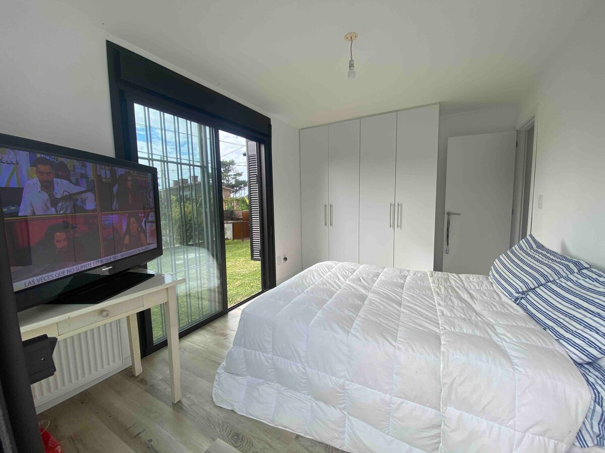 A bright bedroom features a spacious bed covered in a white quilt, with striped pillows adding subtle color. A large window offers views of the garden, while a television stands on a small white desk. Modern cabinetry lines one wall for convenient storage.