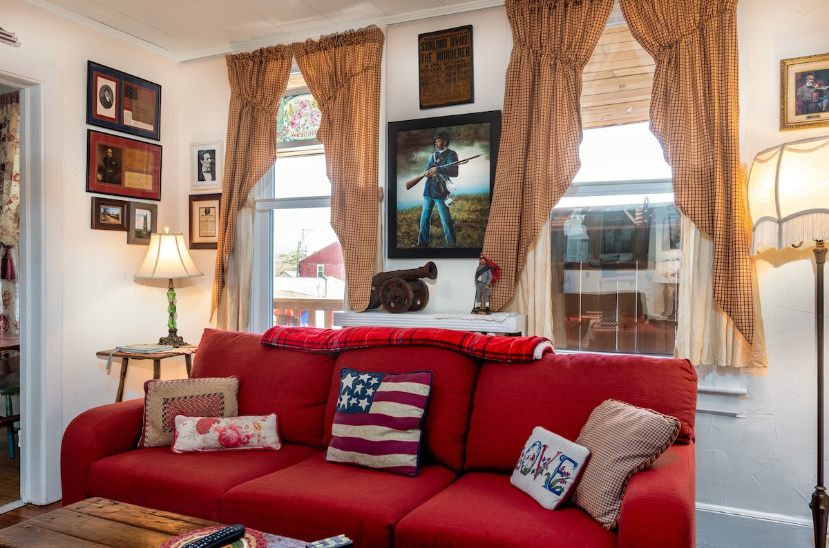A comfortable red sofa is complemented by decorative pillows, including one featuring an American flag. Vintage photos adorn the walls, alongside a large framed image of a soldier. Soft light from a table lamp illuminates the cozy living space, accented by warm curtains framing the windows.