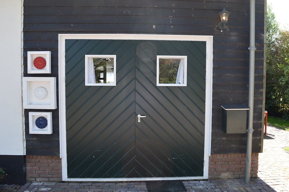 A green, diagonal patterned wooden door is framed by two small square windows. The entryway is flanked by three decorative wall elements featuring circular designs in red and blue. A pathway composed of cobblestones leads to the door, enhancing accessibility.
