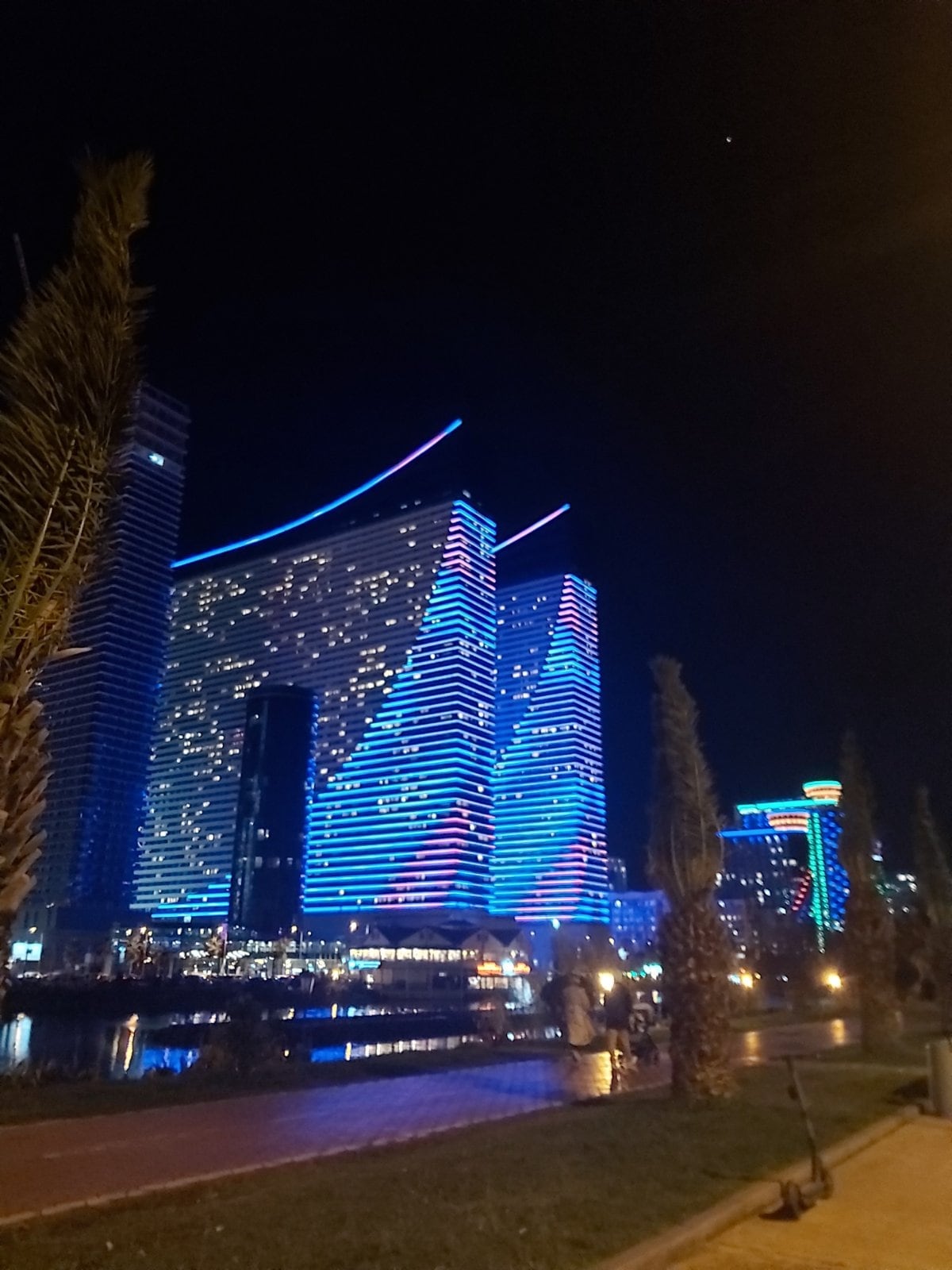 A modern high-rise building is illuminated with vibrant blue and red lights against a dark night sky. The colorful façade reflects in the water below, while palm trees line the pathway, creating a dynamic urban scene.