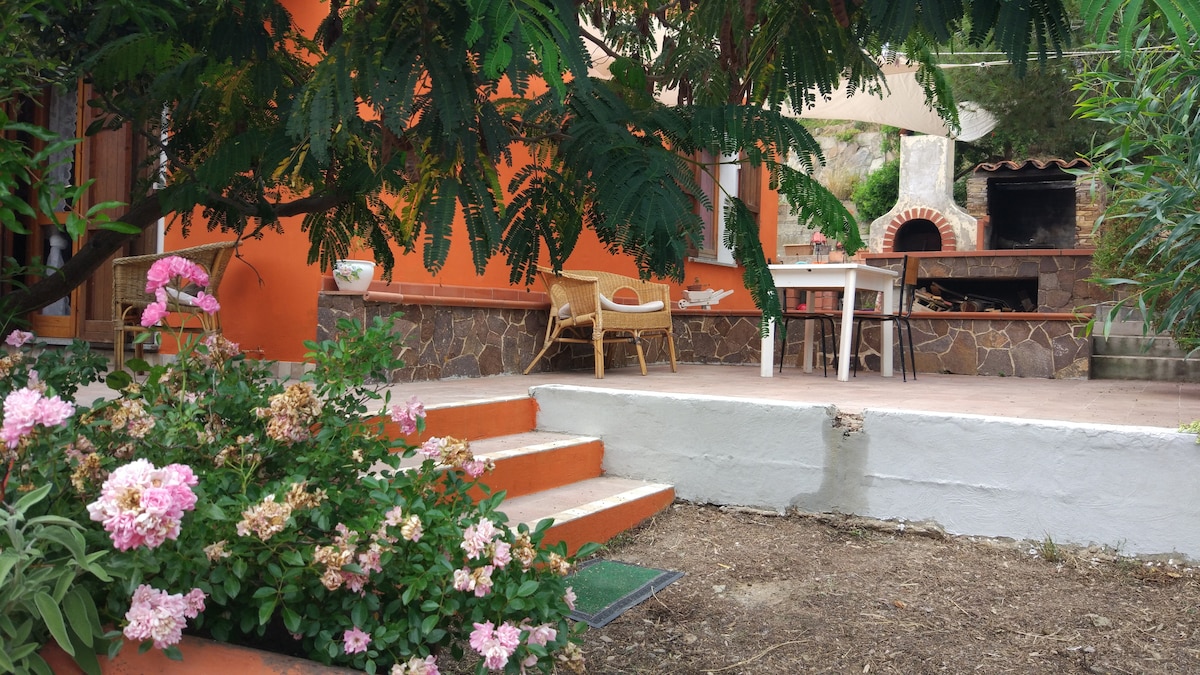 An outdoor area features a stone barbecue and a wood-fired oven, surrounded by vibrant flowers and greenery. A small table with chairs is positioned nearby, offering a space for outdoor dining. The walls of the house are painted in warm orange tones, enhancing the inviting setting.