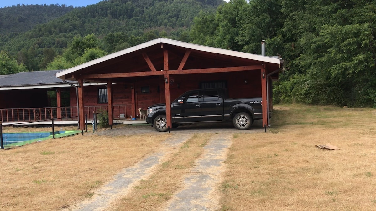 A wooden house situated amidst native forest features a spacious porch supported by large wooden beams. A black pickup truck is parked in the driveway, which leads through dry grass towards the entrance. Forested hills rise in the background, adding to the natural setting.