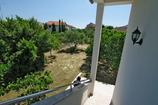 A view from the balcony offers a glimpse of a well-maintained garden, featuring a variety of green trees and foliage. A pathway is visible, leading through the lush space, while a light fixture is positioned on the wall nearby.