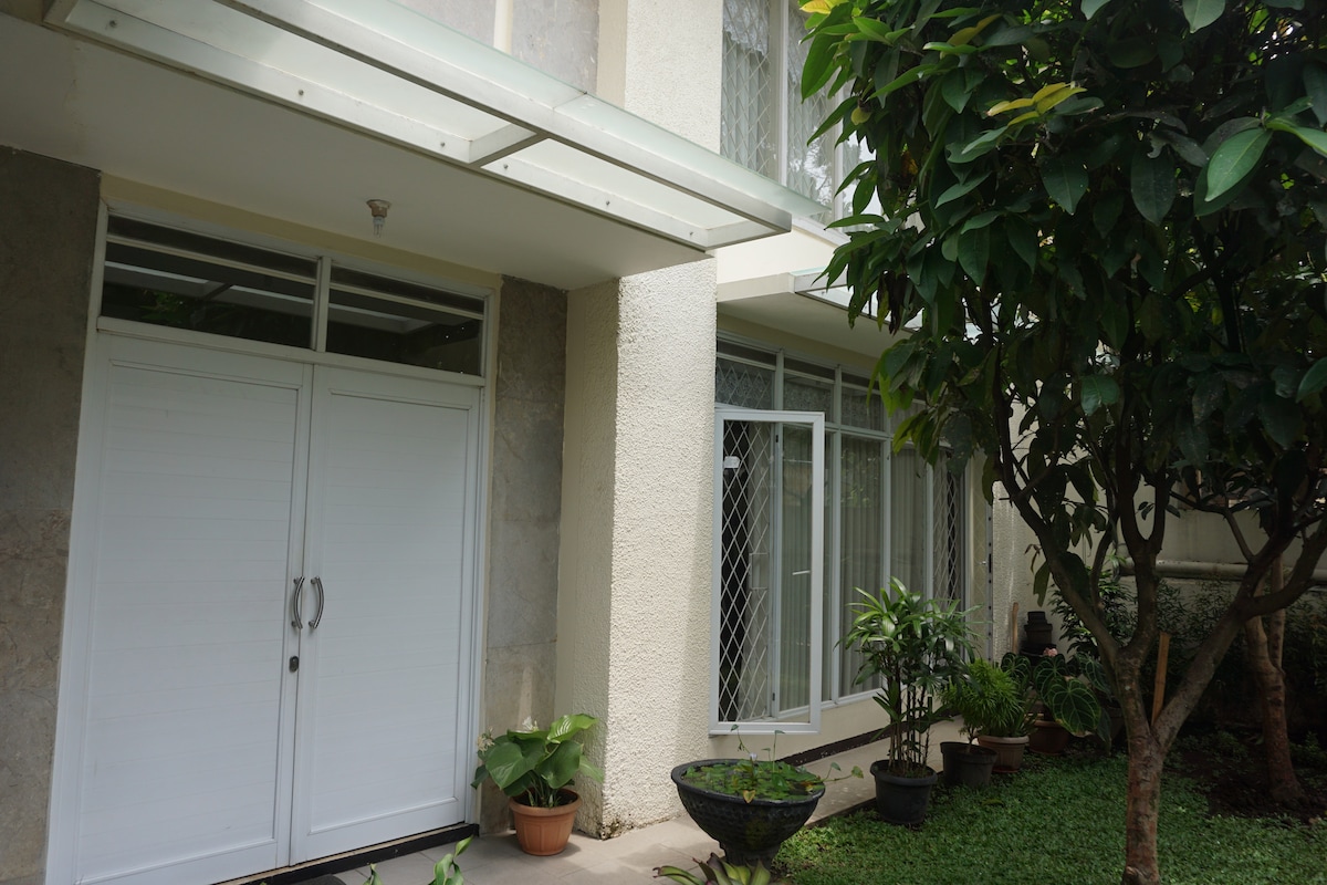 The exterior entrance of the house features large double doors with a white finish, framed by greenery and potted plants. The porch is adorned with a decorative bowl and lush grass, creating an inviting approach to the home.