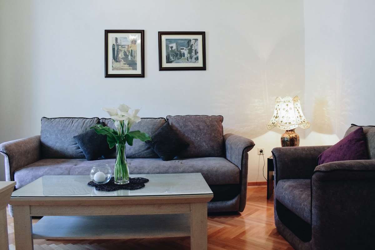 A comfortable seating area features a plush gray sofa and an armchair, complemented by decorative pillows. A coffee table sits in the center, adorned with a clear bowl and a floral arrangement. Soft lighting from a patterned lamp adds a gentle glow to the space.