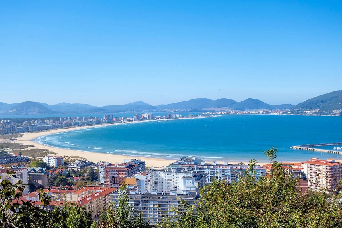 A wide view of the coastline reveals a sandy beach embracing a sweeping bay. The blue water sparkles under clear skies, with distant mountains forming a tranquil backdrop. Urban buildings line the shore, showcasing a variety of architecture, while greenery frames the scene in the foreground.