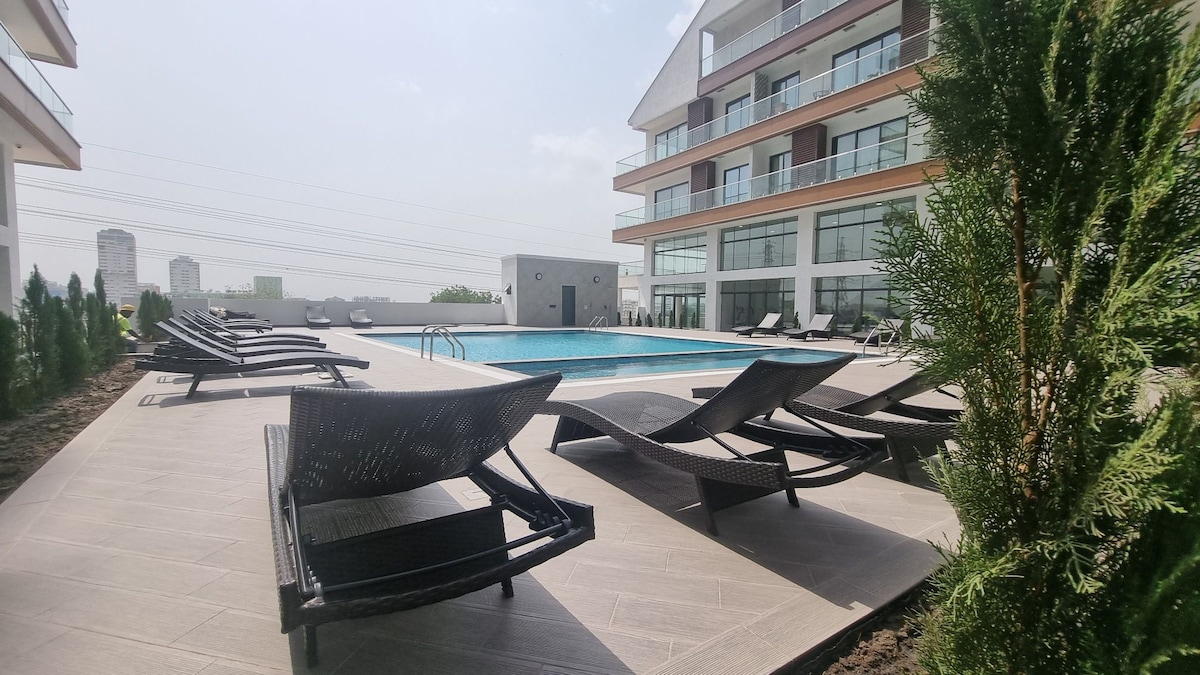 An outdoor pool area is surrounded by lounge chairs arranged neatly on a tiled patio. Lush greenery is planted along the perimeter, and the building's modern design features large glass windows, enhancing the contemporary feel of the space.
