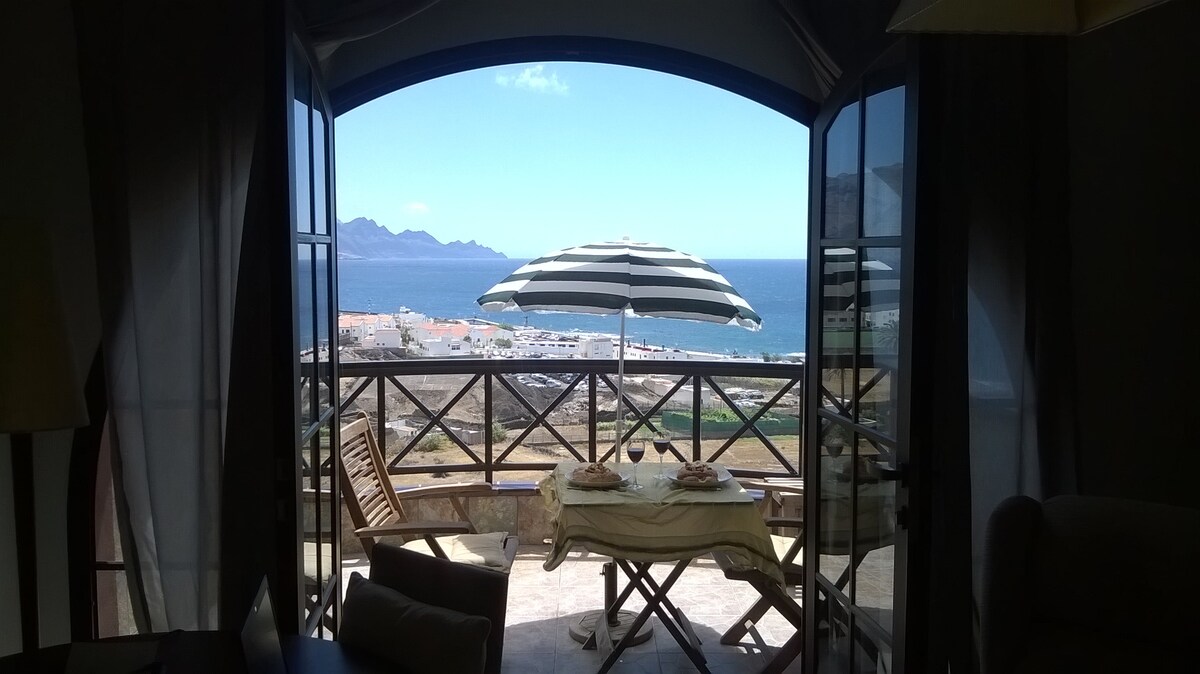 A spacious terrace is visible through open glass doors, featuring a striped umbrella over a dining table set for two. The tranquil sea and distant mountains create a scenic backdrop, complemented by a comfortable seating area nearby.