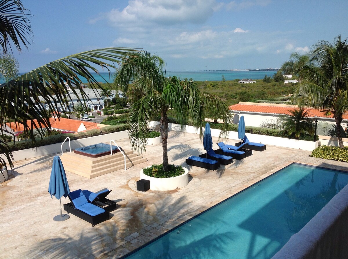 A tranquil pool area features two swimming pools, one heated, surrounded by lush greenery and palm trees. Several blue lounge chairs are positioned under large umbrellas, offering a serene view of the ocean beyond. A hot tub is situated near the edge of the deck.