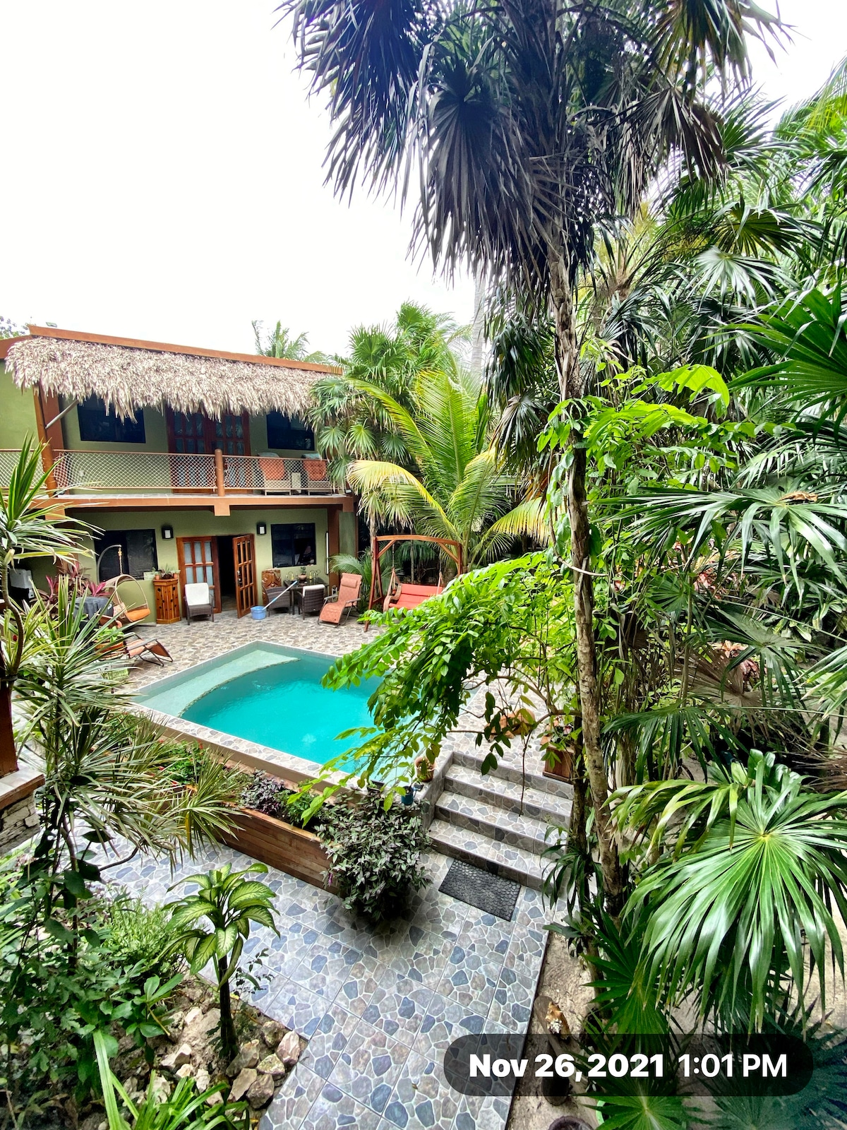 A view of the private courtyard reveals a swimming pool surrounded by tropical plants. Comfortable lounge chairs are positioned next to the pool. A two-story structure with a thatched roof is visible, blending seamlessly with the lush greenery.