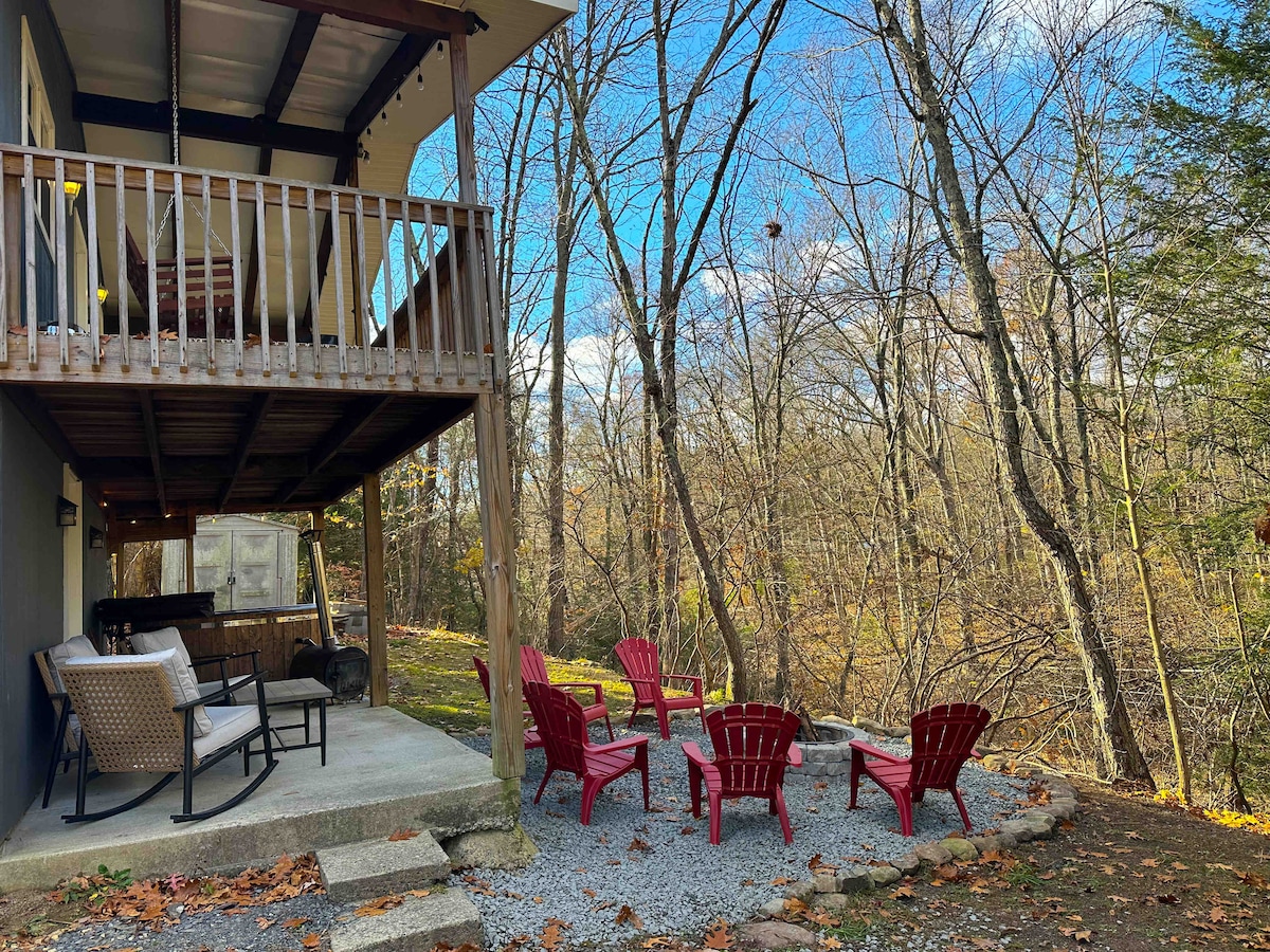 An outdoor seating area features six red Adirondack chairs arranged around a stone fire pit, surrounded by natural landscaping. A covered porch above provides shaded seating options with two woven chairs. Leafless trees and a blue sky create a serene backdrop.