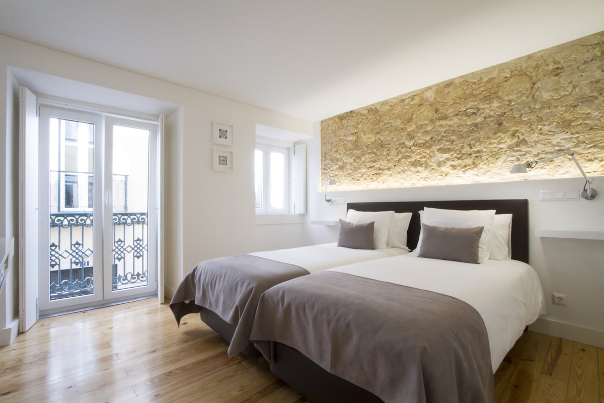 A well-lit bedroom features two comfortable single beds arranged with neutral linens. Natural light enters through traditional windows, highlighting the stone accent wall behind the beds. Minimalistic decor is complemented by modern bedside reading lights, creating a serene and stylish sleeping area.