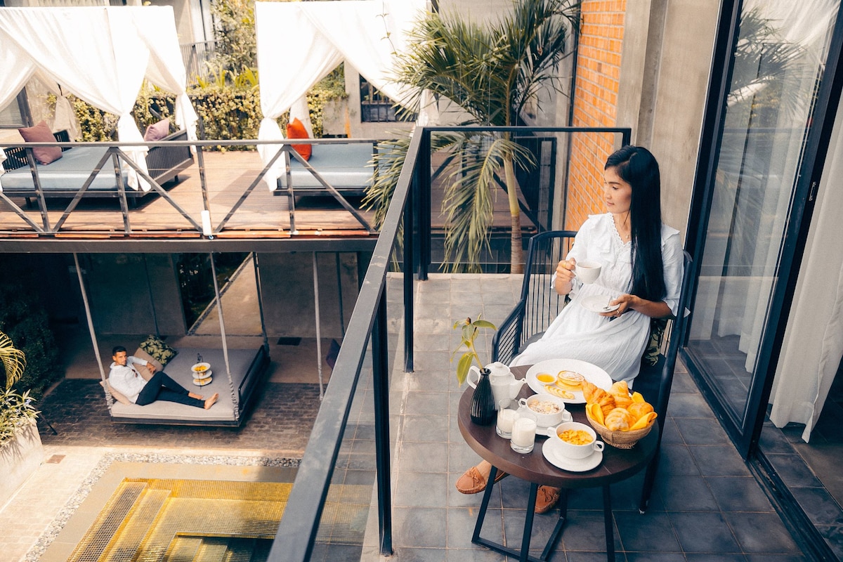 A private terrace offers a table set with a variety of breakfast items, including fruit and drinks. Below, a spacious lounging area features sunbeds under draped canopies. The surrounding greenery and open layout enhance the serene outdoor space.