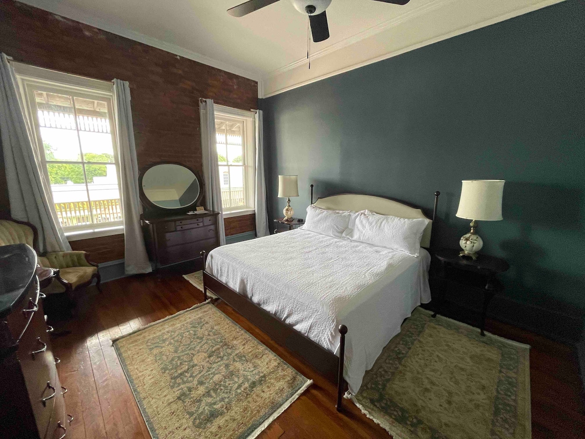 A spacious bedroom features a large bed dressed in white linens. Antique furnishings, including a dresser and bedside tables, are present. Natural light fills the room through two large windows with sheer curtains, highlighting the rich wooden floor and the dark green accent wall.