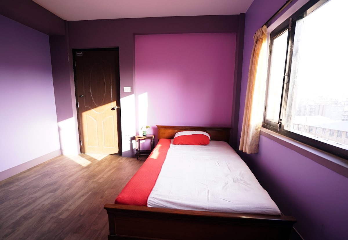 A bedroom features a wooden bed with a vibrant red and white bedding arrangement. Soft natural light filters in through a window, illuminating the lavender walls. A small bedside table is positioned next to the bed, with a green lamp adding a touch of color.