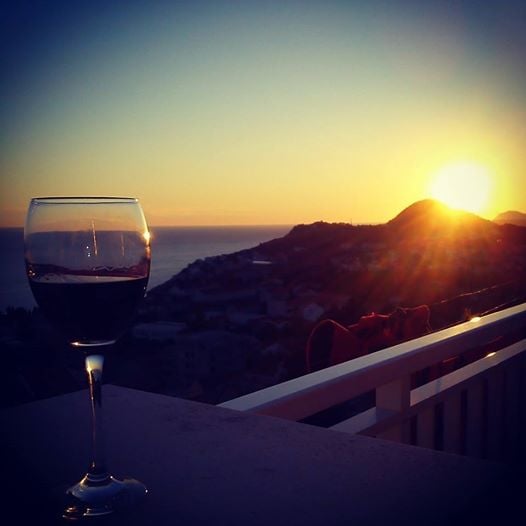 A glass of red wine is placed on a balcony railing, capturing the golden hues of a sunset over the sea. The silhouette of the surrounding hills can be seen in the background, offering a serene atmosphere.