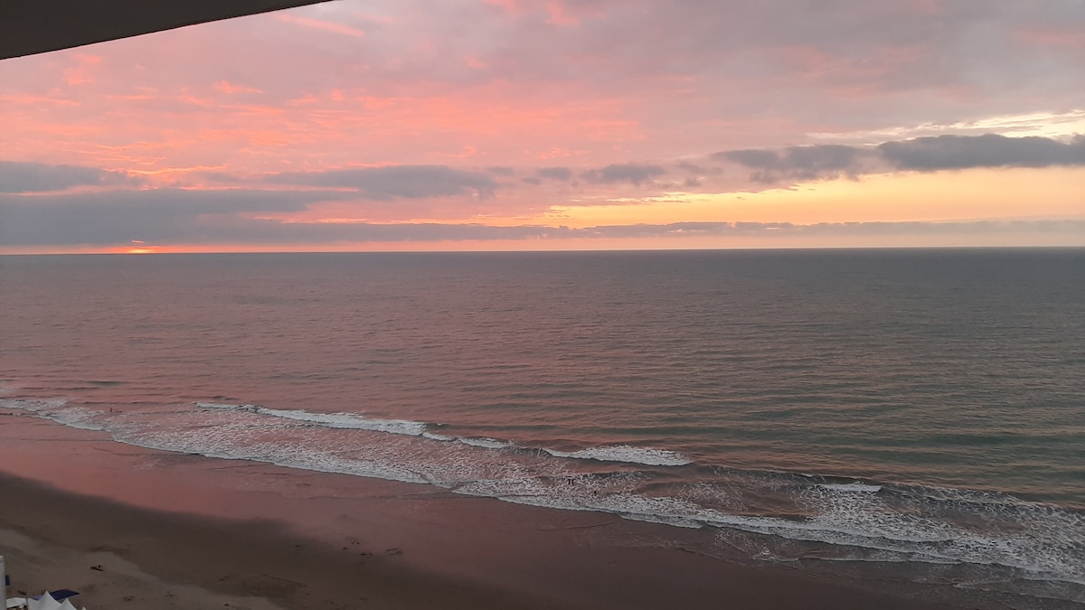 A serene ocean view is presented at sunset, with soft hues of pink, orange, and purple reflecting off the water. Gentle waves lap at the sandy shore, creating a tranquil setting that captures the beauty of the coastline.