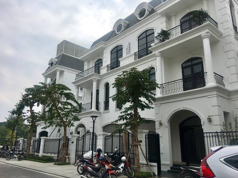 A modern multi-story building is showcased with white exterior walls and architectural details. Balconies with black railings are seen on the upper floors. Lush trees line the front, and parked vehicles are visible along the street, highlighting the property's accessibility.