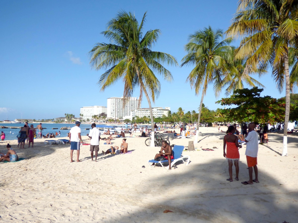 A sandy beach scene bustling with people enjoying the warm weather. Several palm trees provide shade, while lounge chairs are positioned across the sand. Clear blue skies and calm waters contribute to a vibrant atmosphere, with groups of individuals engaged in conversation and leisure activities.